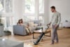 Smiling man vacuums mess from wood floor while female familiy members watch in delight.