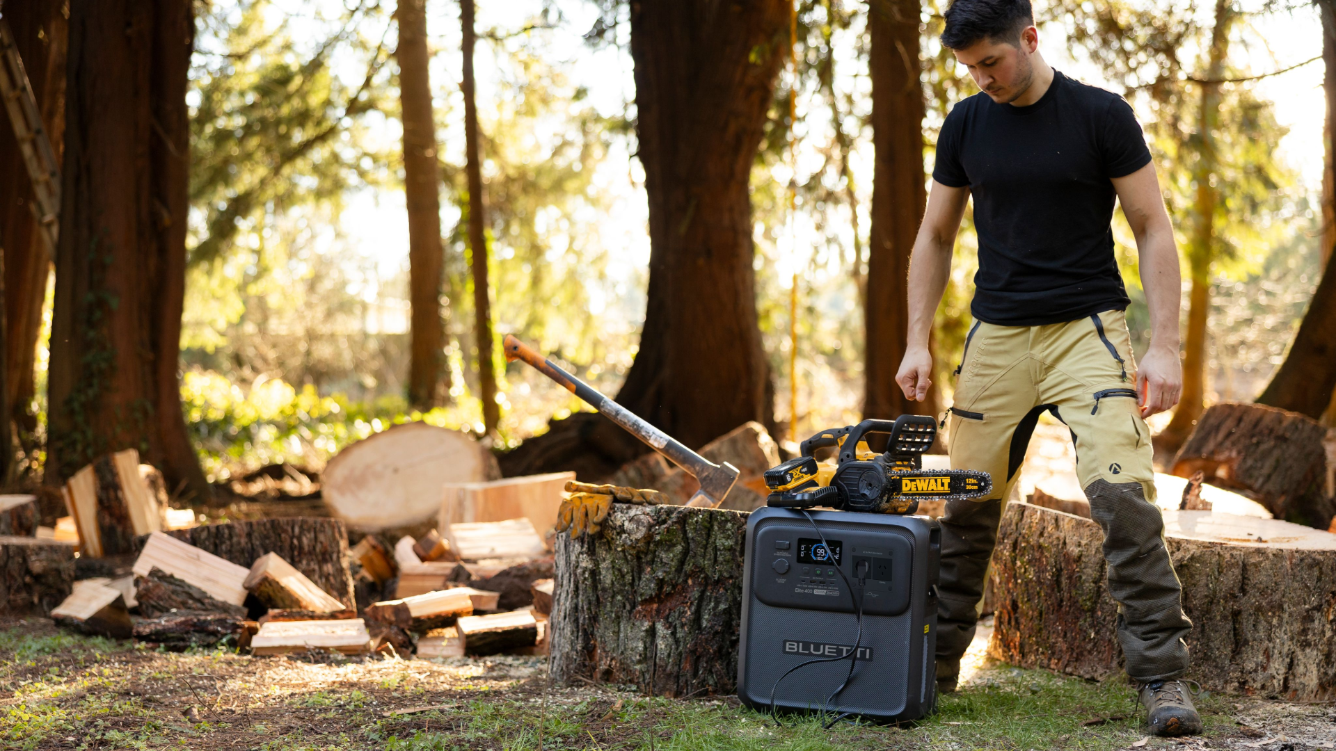 Man at forested worksite with axe, cut timber, trees and power saw being charged by portable power station.