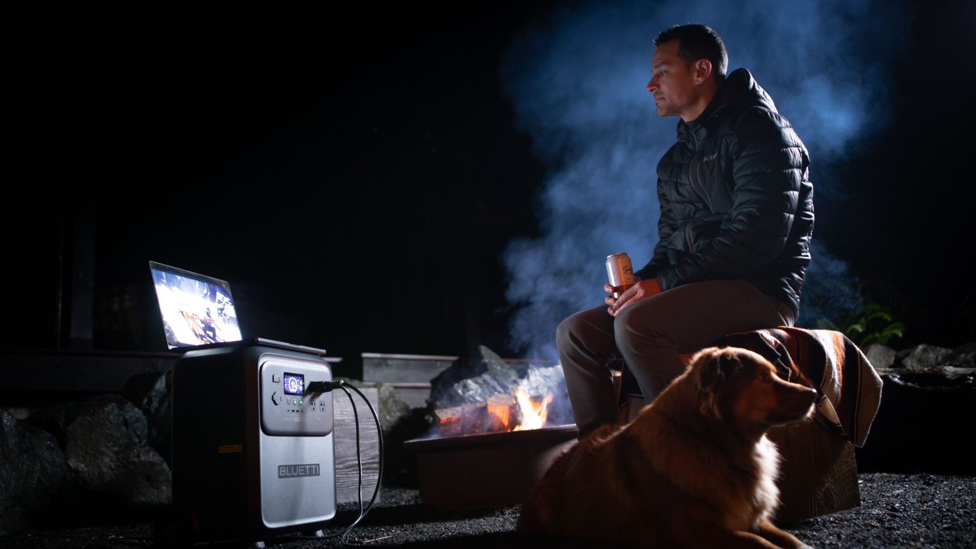 Man at campsite at night with dog and fire, watching sports on a laptop plugged into a portable power supply.